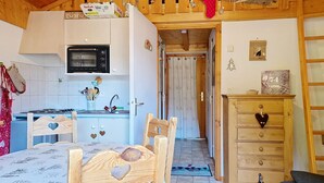 A compact kitchen with a microwave, stove, and wooden cabinets. A dining table with heart-shaped chairs is in the foreground. - Forsythia FA18 FAMILY 1& NATURE 4 Pers. (Châtel)