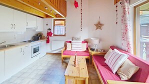A cozy room with a wooden ceiling, a kitchenette, a sofa with a pink cushion, a wooden table, and a window with curtains. - Forsythia FA29 FAMILY 1& NATURE 8 Pers. (Châtel)