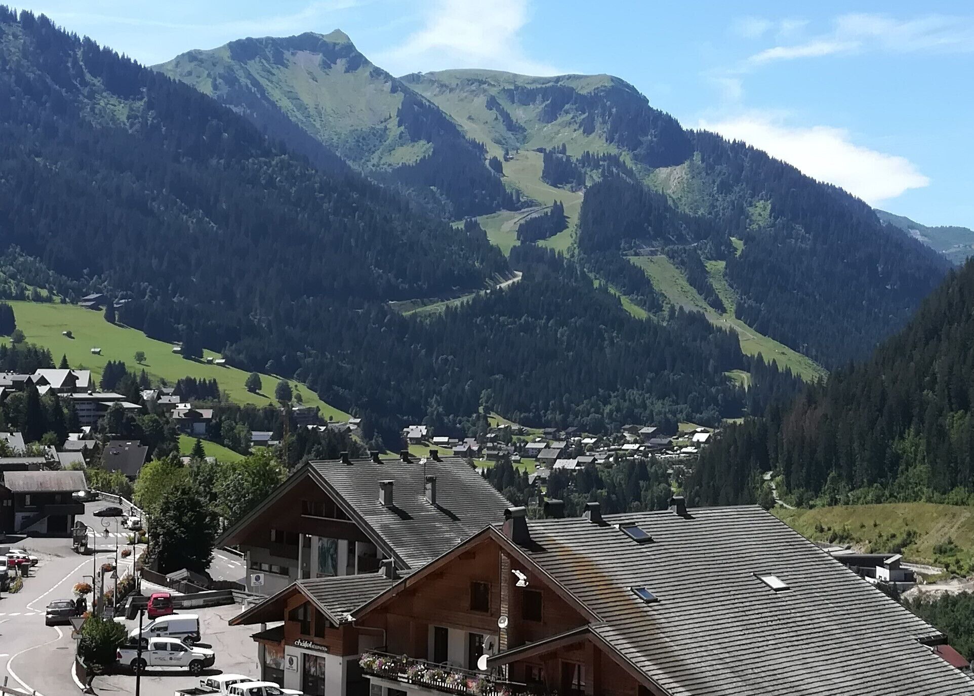 A mountain village with traditional wooden buildings, a main road, and a valley with dense forests and mountains in the background.