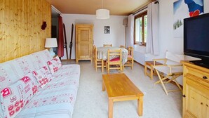 Una sala de estar acogedora con un sofá de madera, una mesa de comedor con sillas, una mesa de centro de madera, un televisor y una ventana con cortinas.
