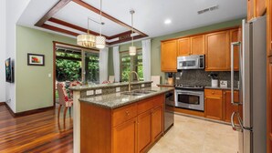 A modern kitchen with wooden cabinets, granite countertops, stainless steel appliances, and a dining area with a view of greenery. - Includes Mauna Lani Beach Club. Fairways at Mauna Lani 201 (Mauna Lani Resort)