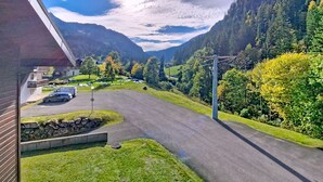 Une vue d’une route menant à une vallée, avec un bâtiment sur la gauche et une colline boisée en arrière-plan.