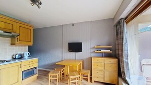 A compact kitchen with wooden cabinets, a built-in microwave, a kettle, and a small table with chairs. A flat-screen TV is mounted on the wall above the table. - SOLARIUM SR19 SUNNY & VIEW 2 Pers. (Châtel)