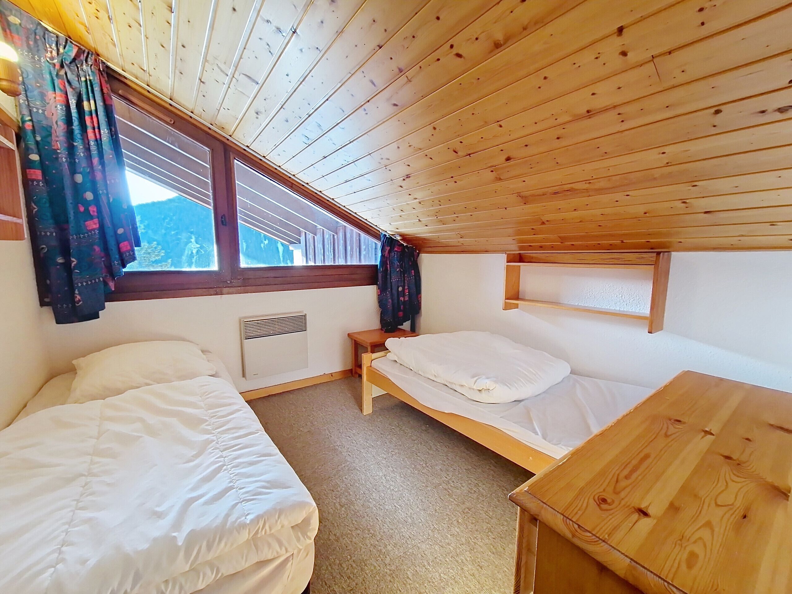 Ein kleiner, gemütlicher Raum mit Holzbalkendecken und -wänden, ausgestattet mit zwei Betten, einem kleinen Tisch und einem Fenster mit Vorhängen.