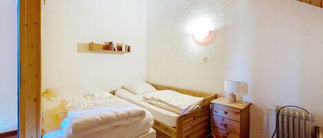 Een kleine, knusse kamer met een houten bed, een nachtkastje met een lamp, een plank met boeken en een radiator.