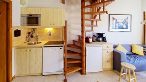 Cocina compacta con armarios de madera, lavavajillas y cafetera. Una escalera espiral de madera conecta la cocina con una sala de estar que tiene un sofá y un cuadro.
