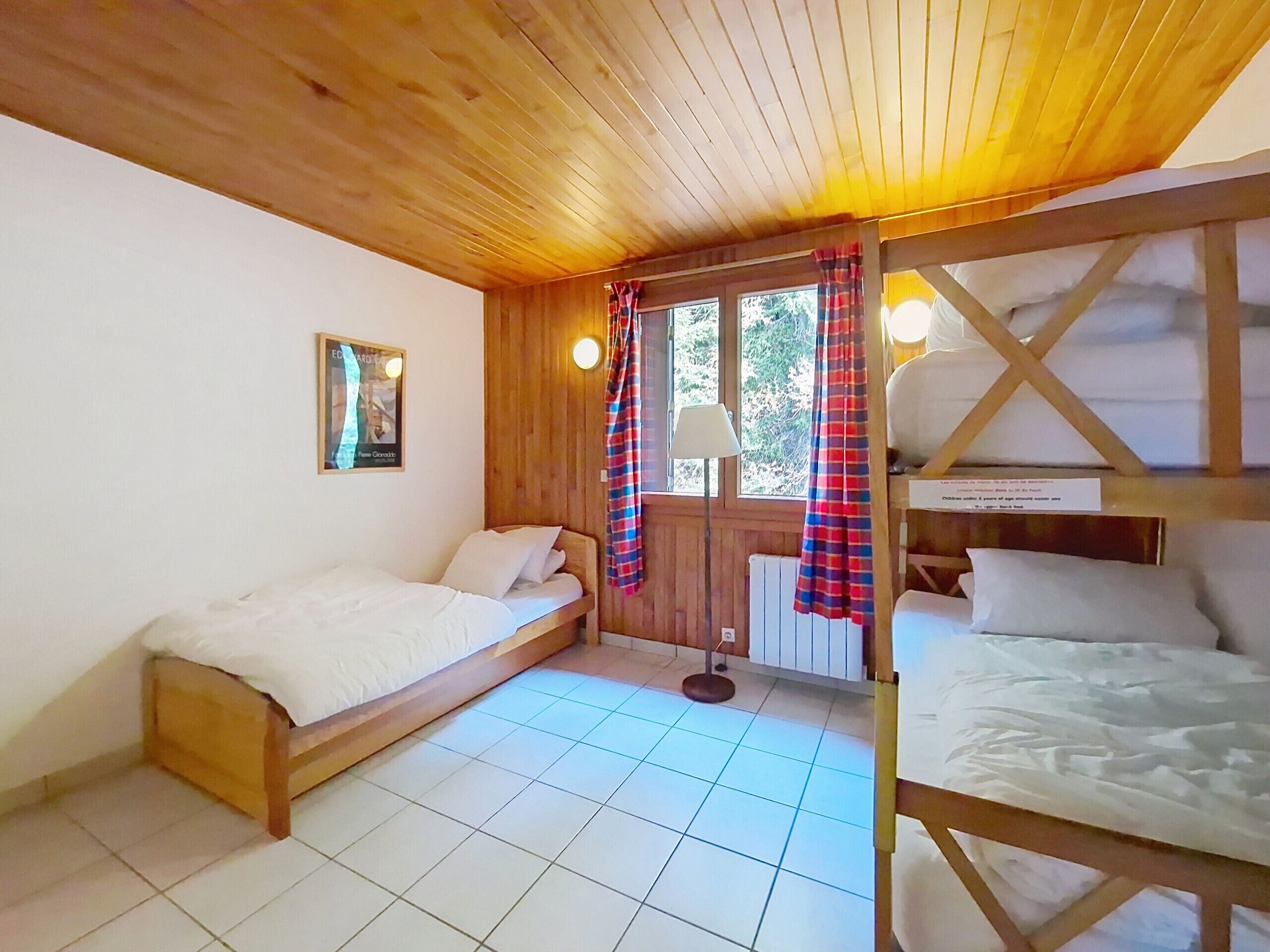 A cozy room with two wooden beds, a window with curtains, a radiator, and a framed picture on the wall.