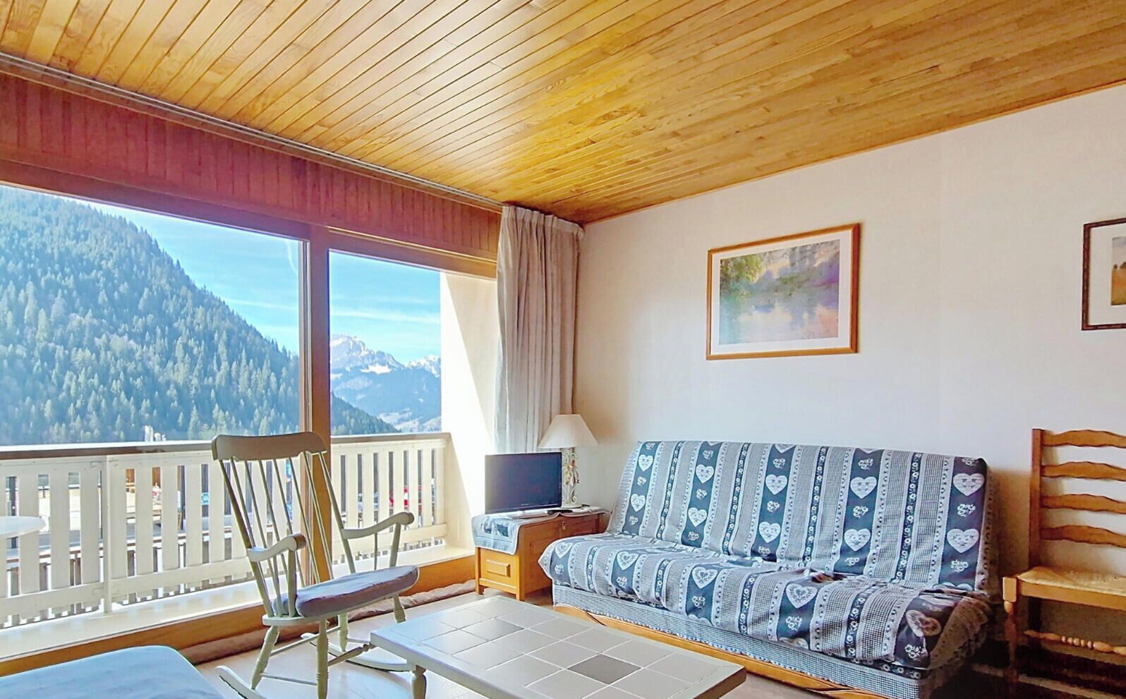 A room with a wooden ceiling, a sofa with patterned cushions, a television, a chair, and a window with a mountain view.