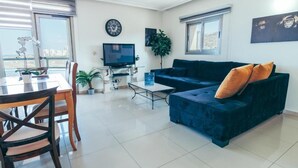 A living room with a dark blue sofa, a dining table with chairs, a TV, and a large window with blinds. - Beautiful Apartments by the sea (Haifa)