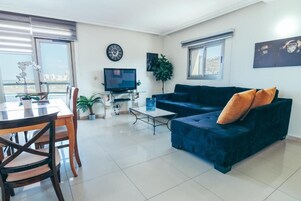 Una sala de estar con un sofá azul oscuro, una mesa de comedor con sillas, un televisor y un ventanal con persianas.