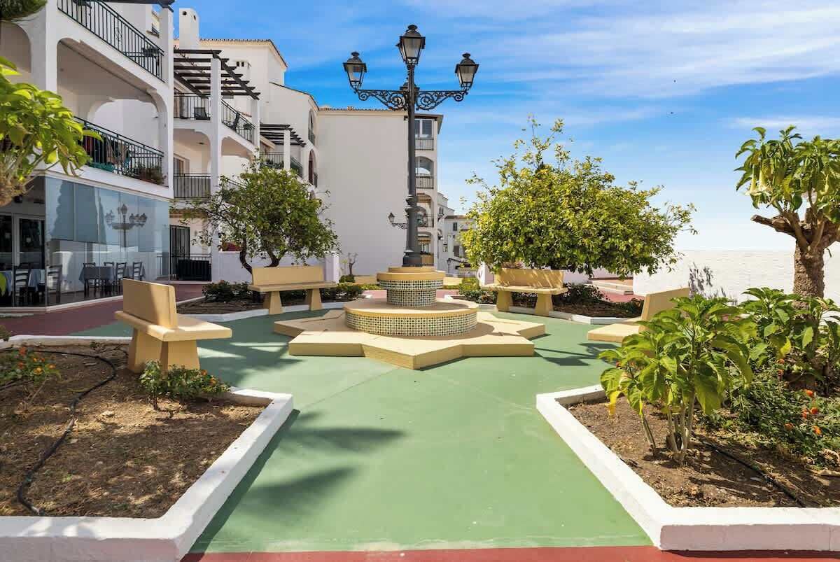 A courtyard with outdoor seating, a central decorative fountain, and a view of the sea.