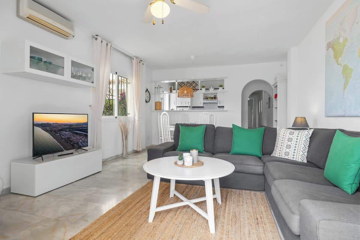 A modern living room with a flat-screen TV, a grey sofa with green cushions, a white coffee table, and a kitchen area in the background.