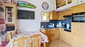 A cozy kitchen with wooden cabinets, a dining table with chairs, a wall-mounted clock, and a flat-screen TV. - Chalet Madeline MD25 SUN & VIEW 6 pers. (Châtel)