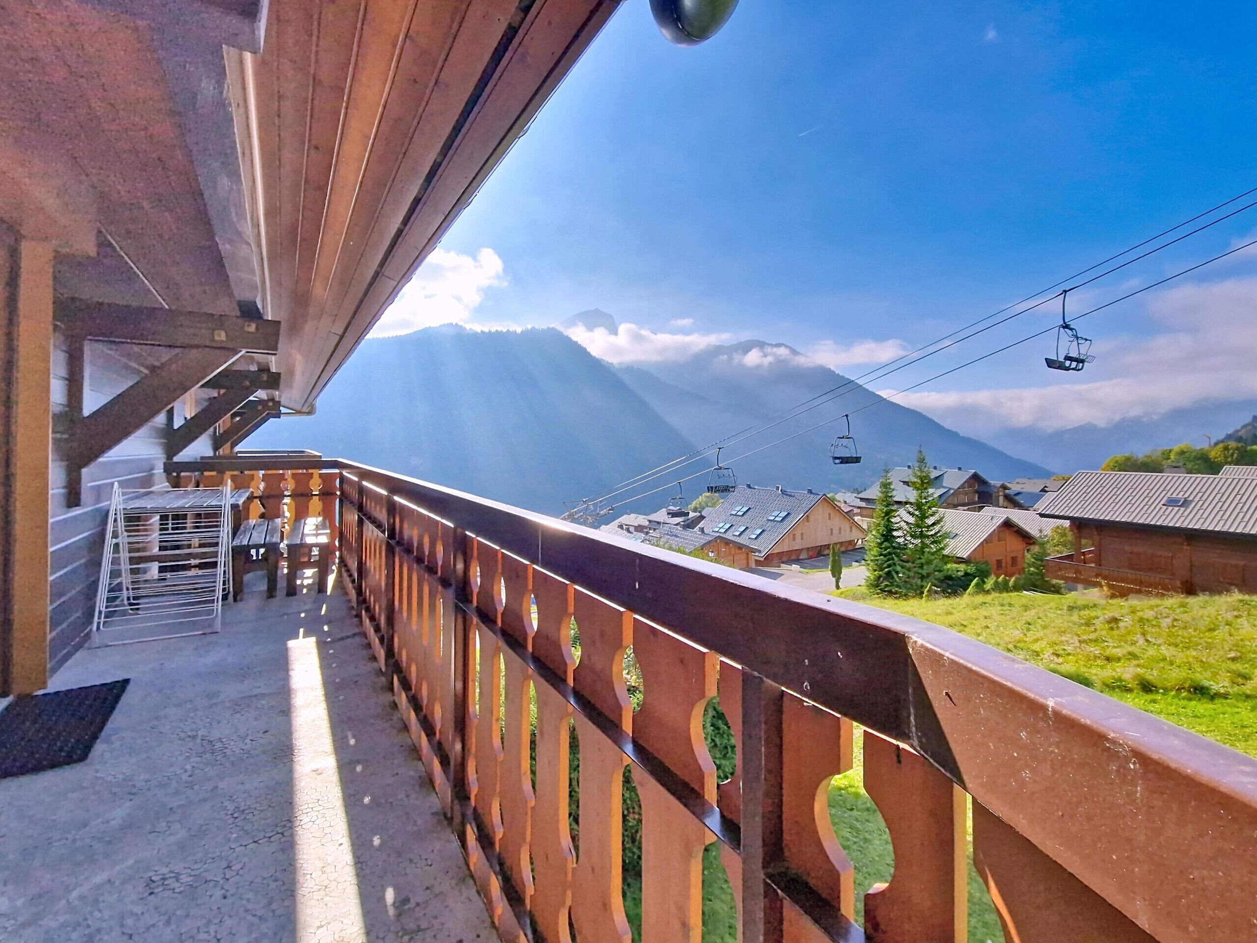 A wooden balcony with a metal railing overlooking a mountainous landscape, a ski lift in the background, and a clear sky.
