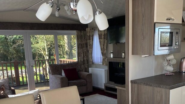 A modern living room with a dining area, a kitchen with a built-in microwave, and a large window offering a view of the outdoors. - Relaxing Lodge In The Heart Of Northumberland (Felton)