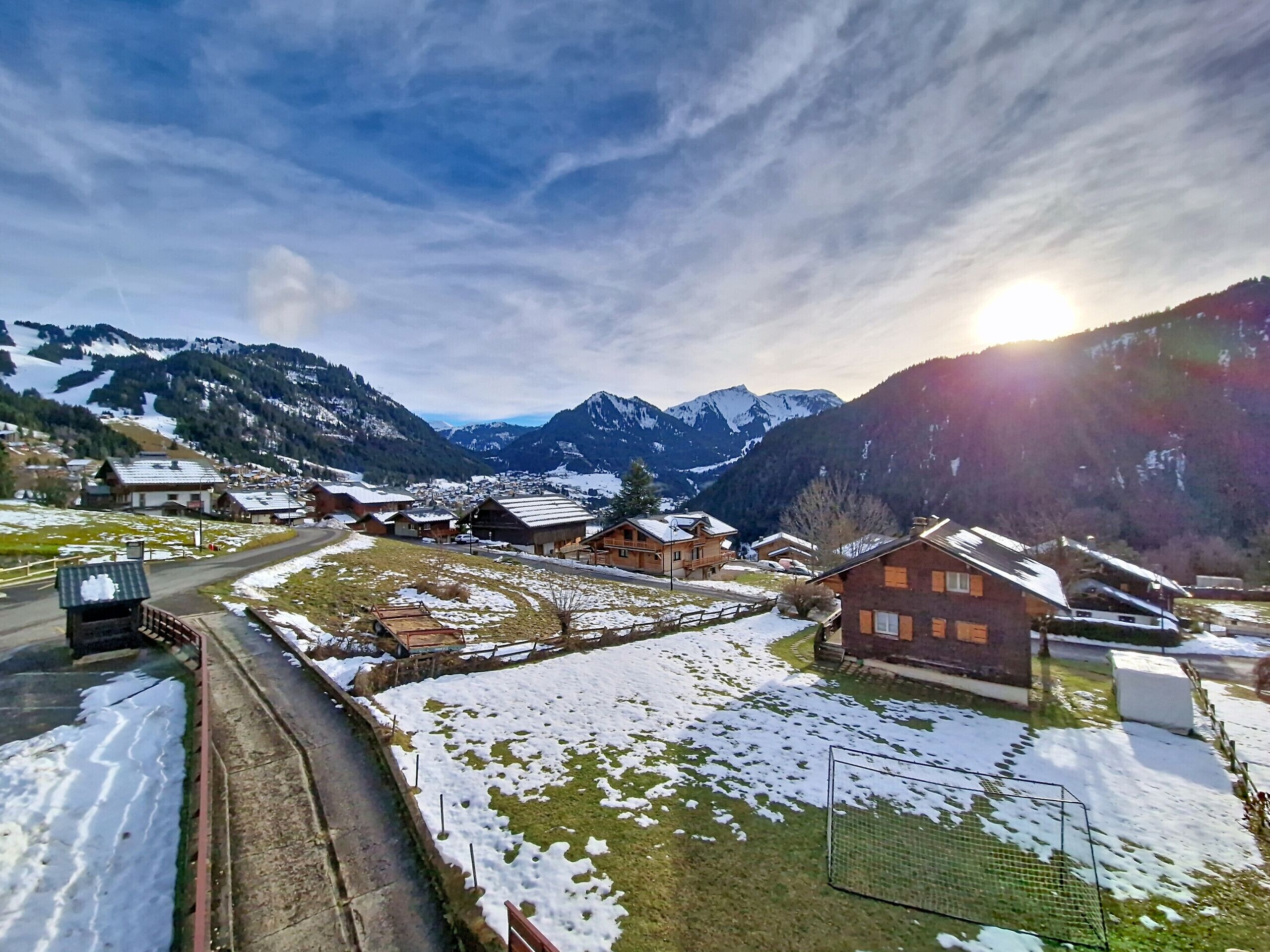 Un pueblo de montaña con casas de madera tradicionales, una carretera y terreno cubierto de nieve.