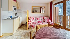 A compact room with a kitchenette, a bed with a patterned cover, a wooden table, and a large window with a view of trees. - Orchidée OD8 NATURE & TELECABINE 4 pers. (Châtel)