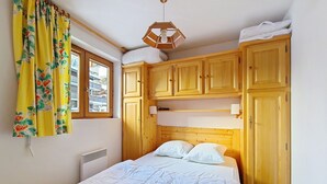 A bedroom with a bed, wooden cabinets, a window with curtains, and a ceiling light fixture. - Orchidée OD8 NATURE & TELECABINE 4 pers. (Châtel)