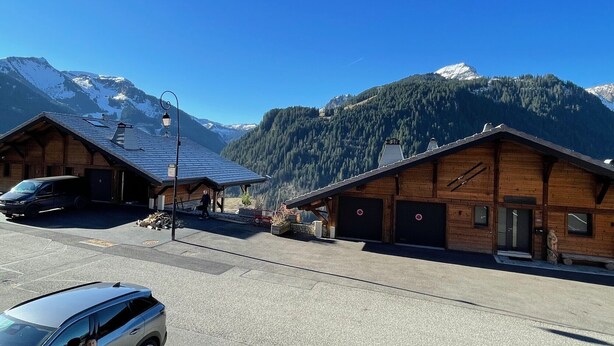 Un refugio de montaña con fachada de madera, una zona de estacionamiento techada y vistas a montañas nevadas.