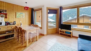 Una habitación acogedora con paredes de madera, una mesa de comedor con sillas, un televisor y ventanas amplias con vistas a la montaña.