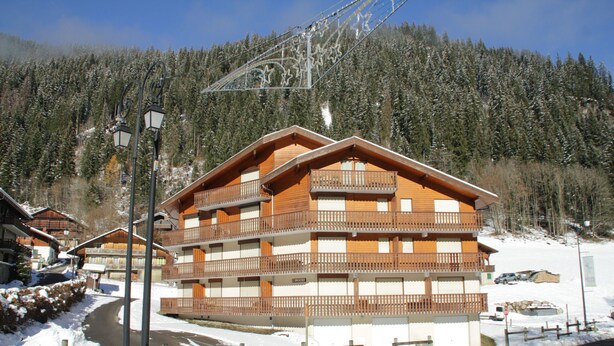 A multi-story wooden building with balconies, surrounded by snow-covered trees and a clear sky. - Orchidée OD5 NATURE & TELECABINE 4 pers. (Châtel)