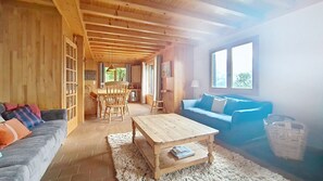 A cozy living room with a wooden ceiling, a blue sofa, a wooden coffee table, and a dining area with a wooden table and chairs.