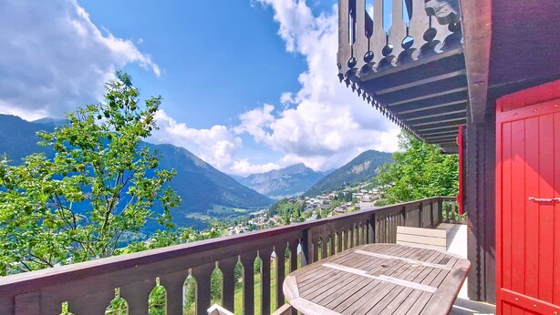 A wooden table and bench on a balcony with a red door, overlooking a mountainous landscape.