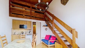 A cozy room with a wooden staircase, a dining table with a patterned tablecloth, a blue sofa with decorative pillows, and a kitchen area with wooden cabinets and a microwave. - Hameau des 4 saisons HSA20  VIEW  & SUN  4 pers. (Châtel)
