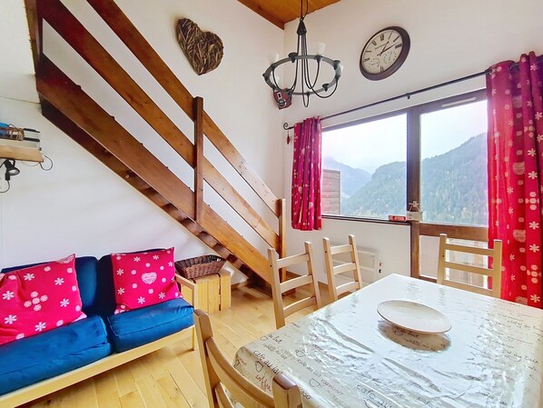 Ein gemütliches Zimmer mit einer Holztreppe, einem Sofa mit gemusterten Kissen, einem Esstisch mit Teller und Bergblick durch ein Fenster.
