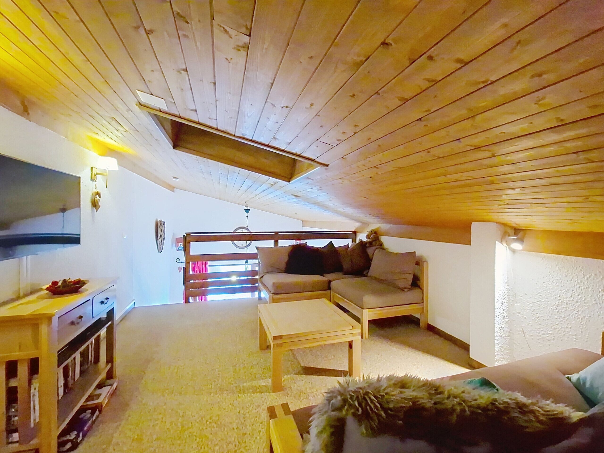 Ett mysigt rum på vinden med ett träpanelat tak, en soffa, ett litet bord och en tv på väggen till vänster.
