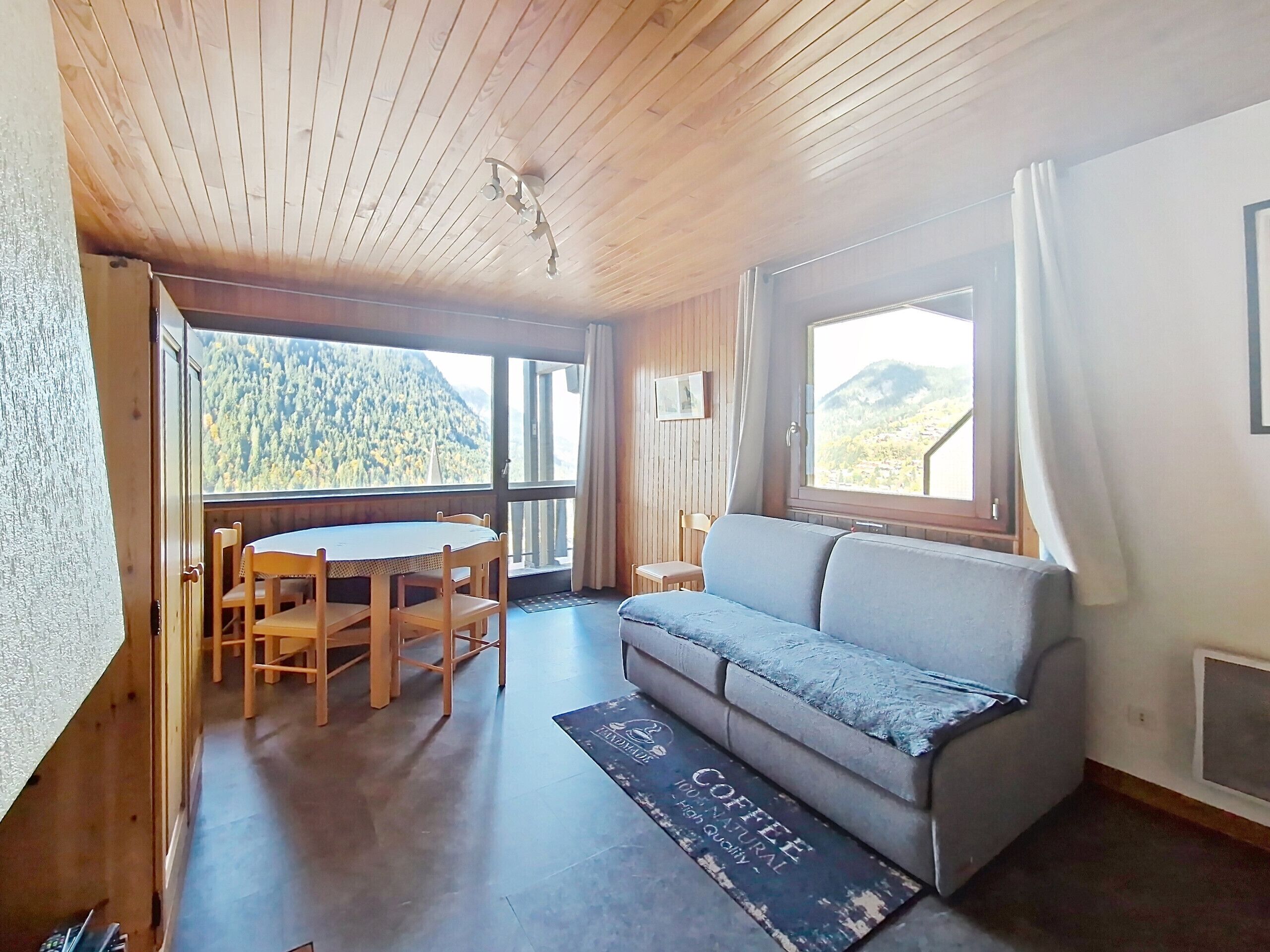 A room with a wooden ceiling, a sofa, a dining table with chairs, and a view of a forested mountain.