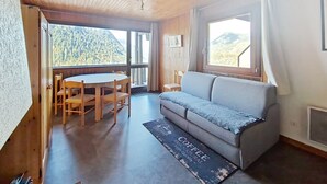 A room with a wooden ceiling, a sofa, a dining table with chairs, and a view of a forested mountain. - Rhododendrons RD501 CENTER & TELECABINE 4 Pers. (Châtel)