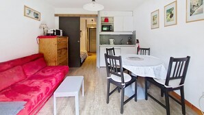 Un espacio habitable compacto con un sofá rojo, una pequeña mesa de comedor con sillas, una kitchenette y un armario de madera.
