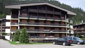 Un edificio de varios pisos con balcones, un estacionamiento y montañas al fondo.