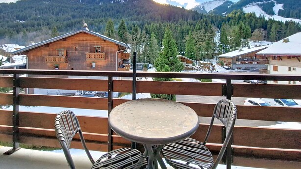 A wooden balcony with a round table and two chairs, overlooking a mountain landscape with buildings and snow. - Portes Du Soleil PSB204 TELECABINE & MOUNTAIN 4 pe (Châtel)