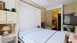 A compact room with a bed, a desk, a chair, a small kitchenette, and a door leading to another room. - Portes Du Soleil PSA46 TELECABINE & MOUNTAIN 4 Per (Châtel)