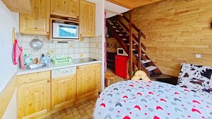 Cocina funcional con armarios de madera, lavavajillas Bosch y microondas en la encimera. Una escalera de madera conduce a un dormitorio con cama cubierta con un edredón estampado.
