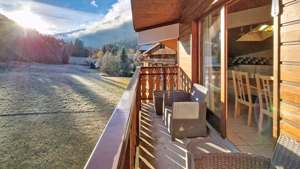 A wooden balcony with wicker furniture, a glass door leading to a dining area, and a view of a snowy landscape with trees and a mountain in the background. - Myrtilles MR27 TELECABINE & NATURE 6 pers. (Châtel)