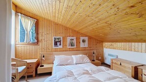 A cozy bedroom with a wooden ceiling, a large bed, a bedside table, a chair, and a window with curtains. - Myrtilles MR27 TELECABINE & NATURE 6 pers. (Châtel)