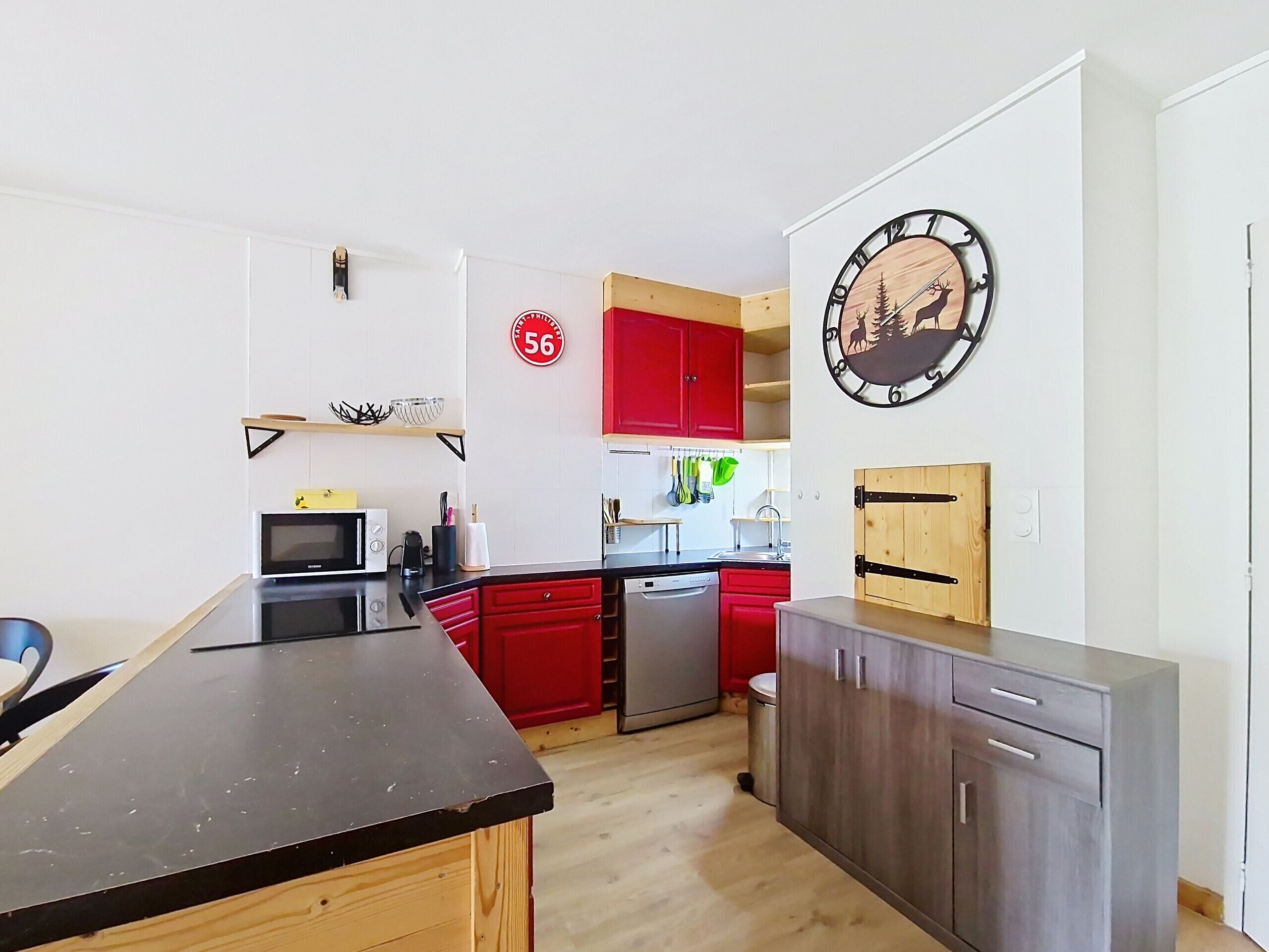 A modern kitchen with red cabinets, a black countertop, and a clock with a deer and trees design.