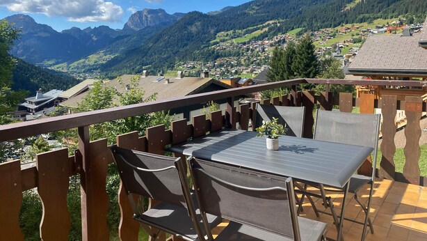 A terrace with a table and chairs, overlooking a mountainous landscape with a village below.