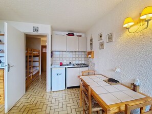 Une cuisine compacte équipée d’une cuisinière, d’un évier et de placards de rangement. Une table à manger avec des chaises est placée devant la cuisine.
