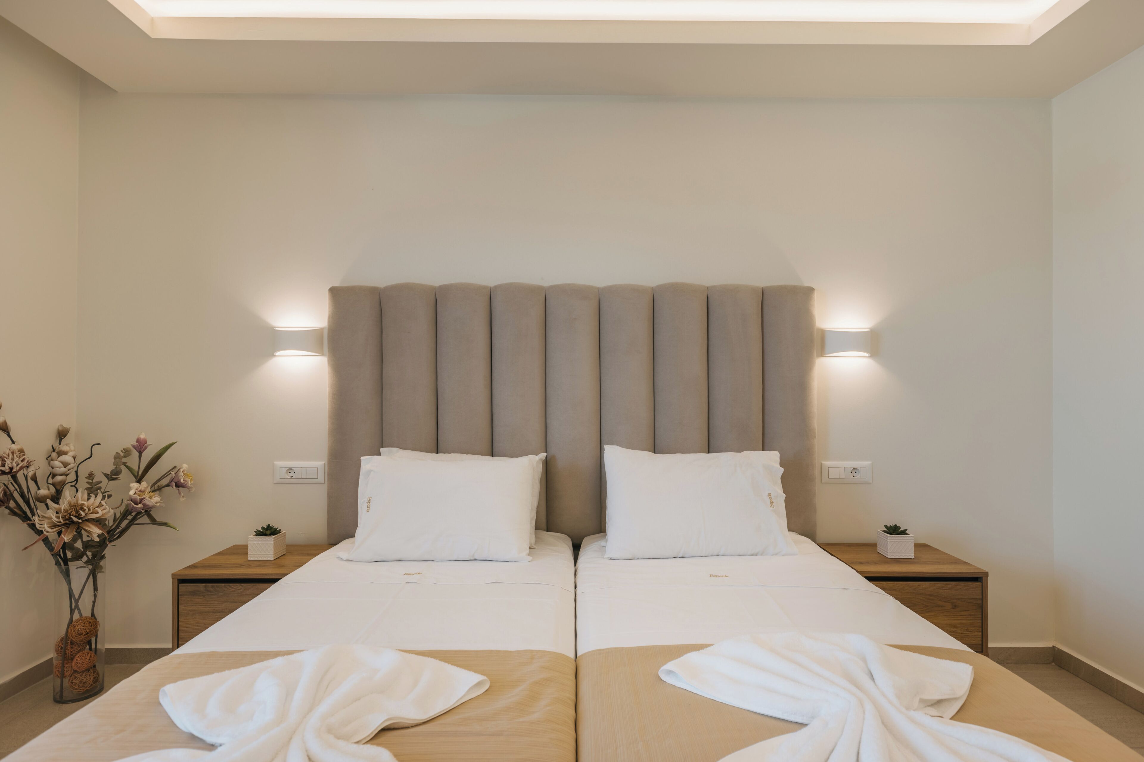 Uma cama de casal com cabeceira, mesas de cabeceira e um vaso com flores.