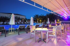 An outdoor pool area with lounge chairs and umbrellas, a covered seating area with tables, and a view of the pool and surrounding landscape.
