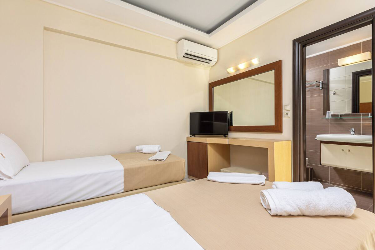 A hotel room with two beds, a TV, a bathroom with a mirror and sink, and an air conditioning unit.