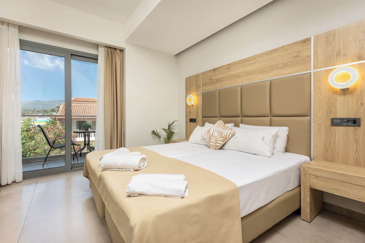 A hotel room with a large bed, a wooden headboard, a nightstand, and a balcony with a view of the landscape.