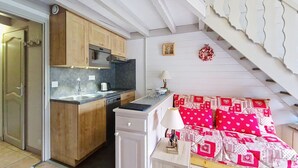 Eine kompakte Küche mit Holzausbau, einem eingebauten Backofen und einer Spüle. Angrenzend an die Küche befindet sich eine kleine Essecke mit Tisch und Stühlen. Eine Treppe mit weißen Geländern führt auf eine obere Ebene.