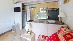 Ein gemütlicher Wohnraum mit Küchenzeile, einer Couch, einem kleinen Tisch mit Lampe und einem an der Wand befestigten Fernseher.