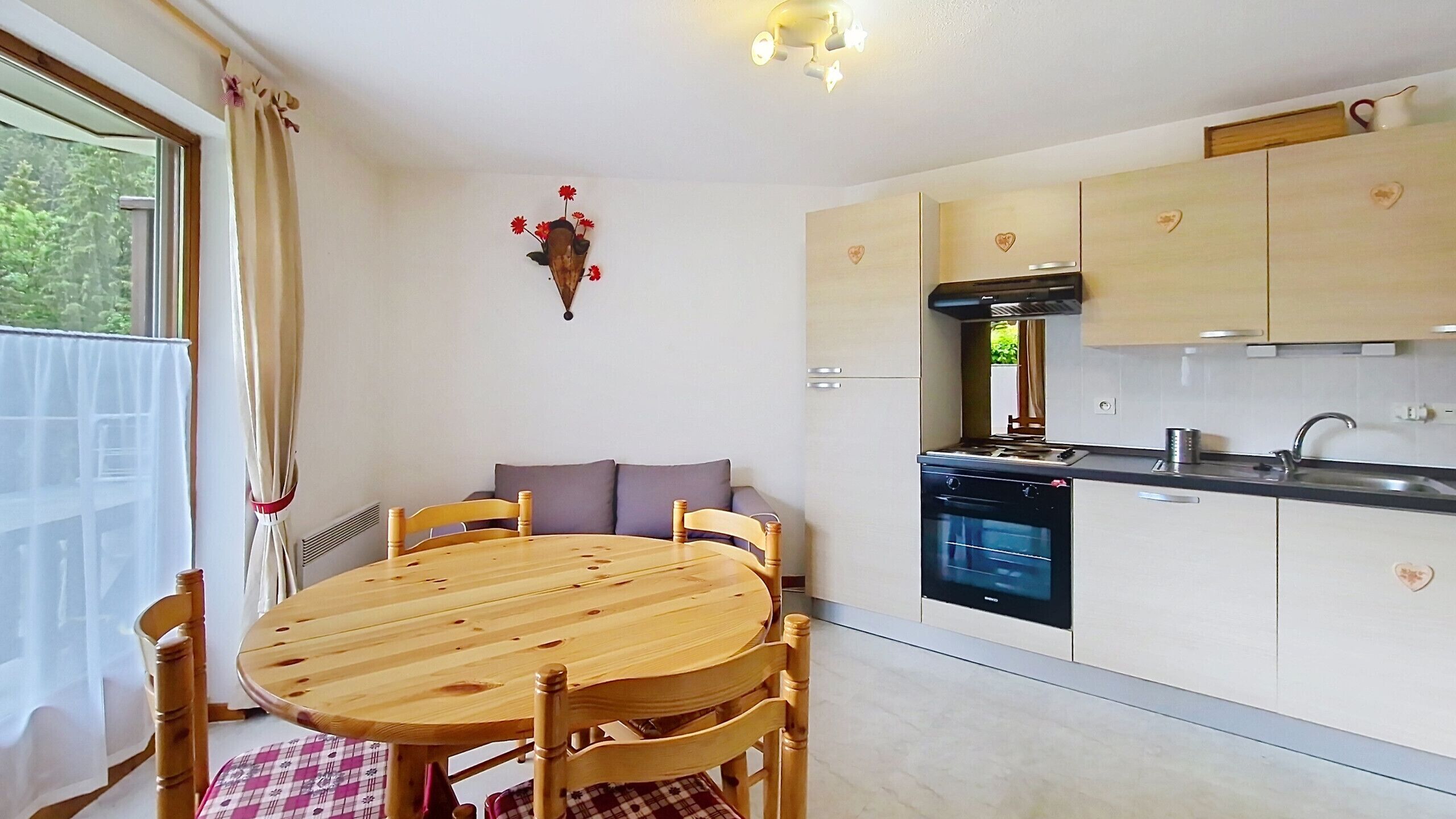 A dining area with a round wooden table and chairs, a kitchen with white cabinets and a stove, and a window with curtains.