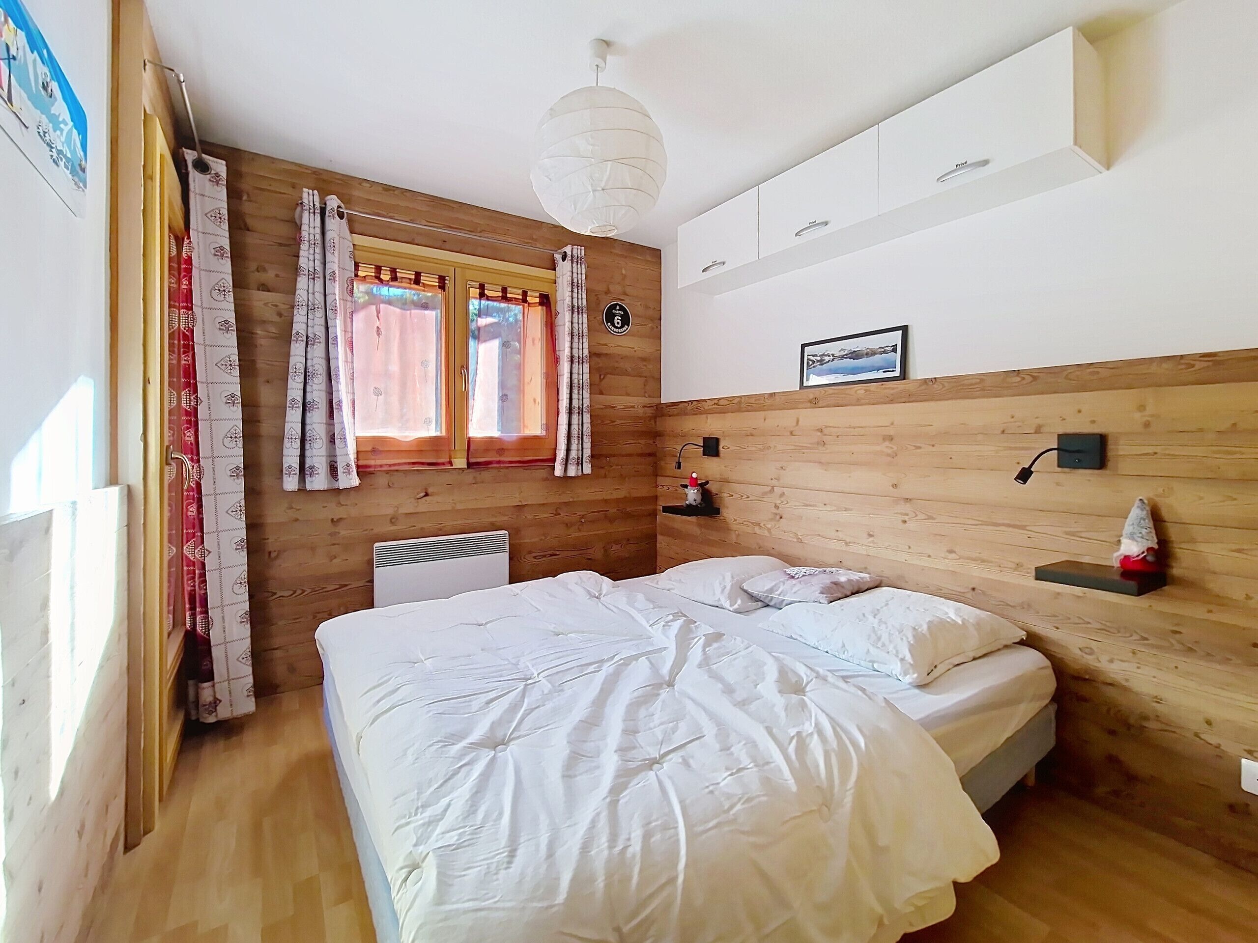 A bedroom with a wooden headboard, a bed with white bedding, a window with curtains, a wall-mounted shelf with a lamp, and a picture frame.
